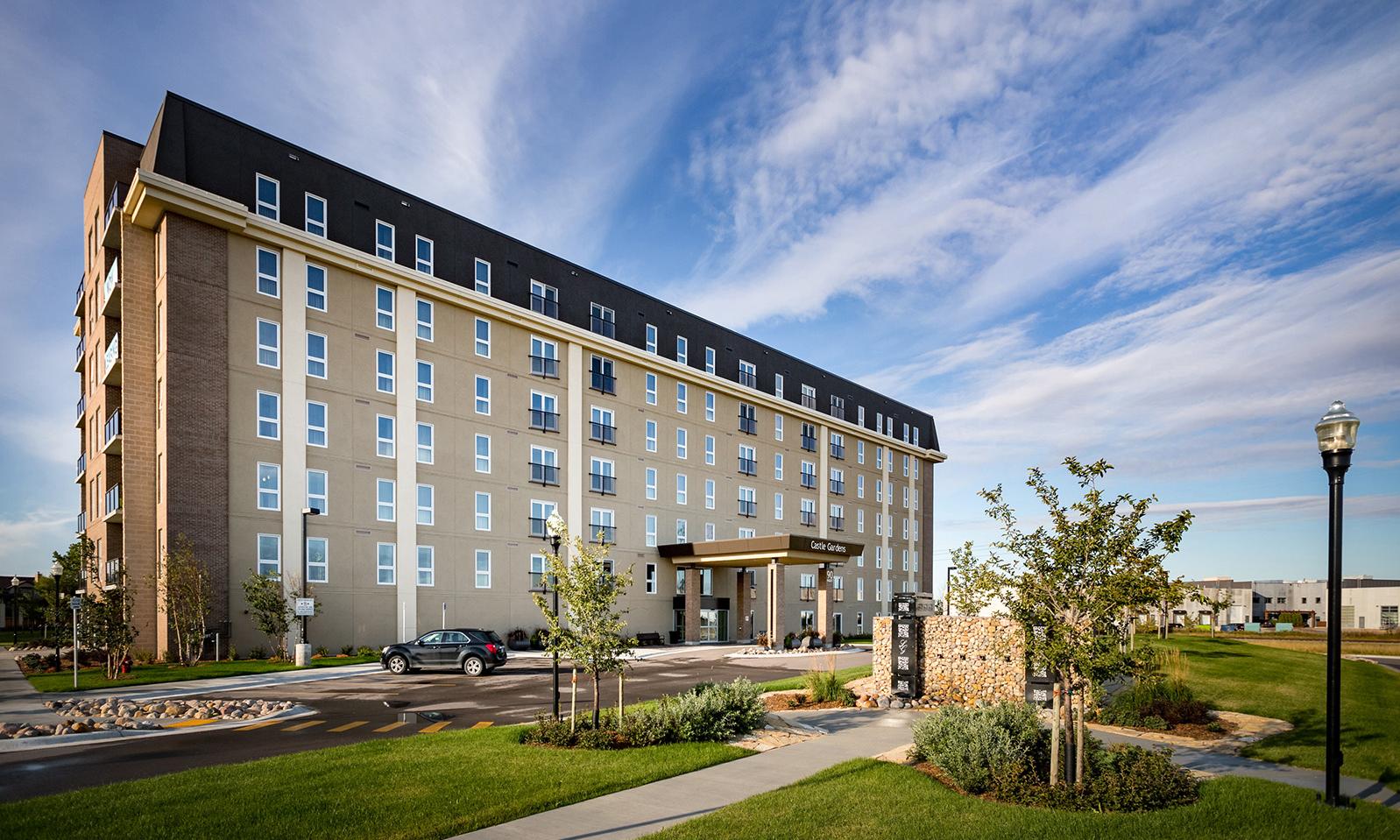 Castle Gardens. An expanse of green lawn with a metal gatepost and small trees and a parking lot. The seven storey apartment building is in the background. 